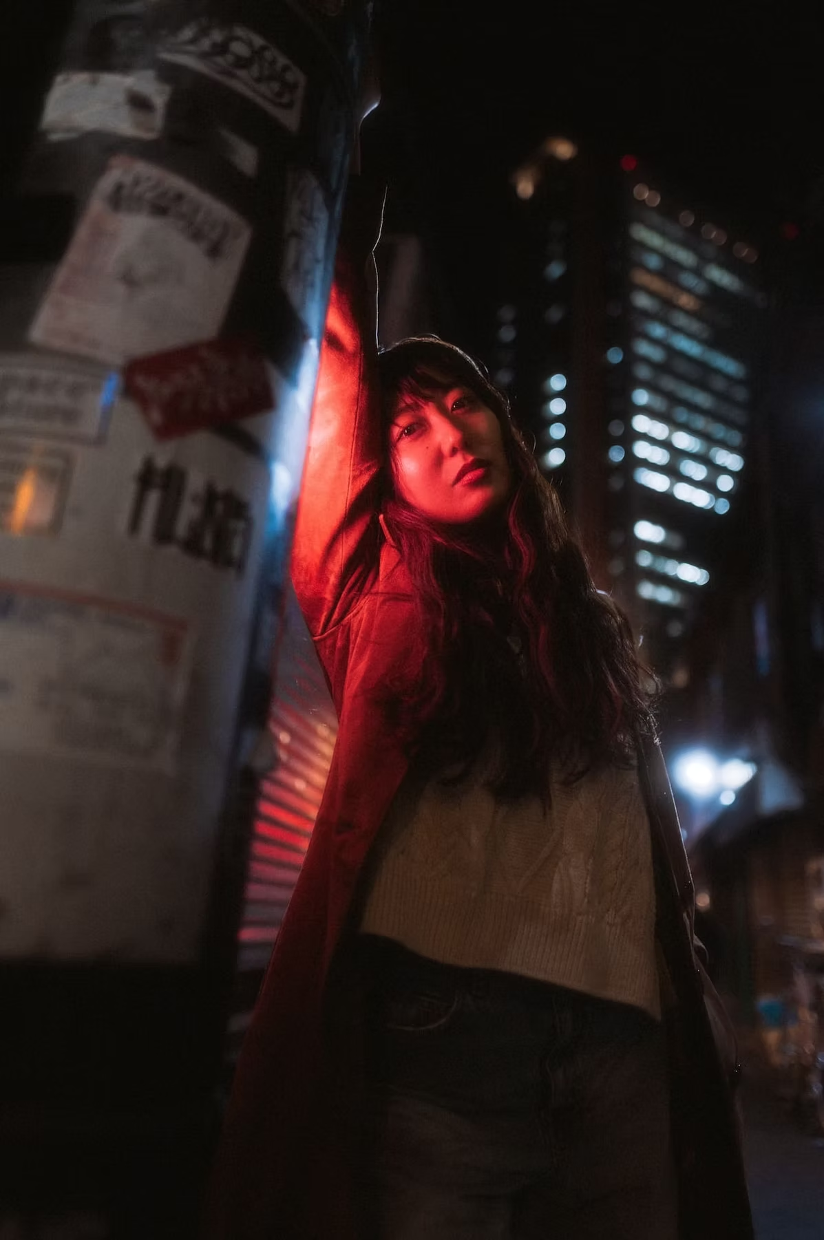 A woman with long hair and a red glow on her face stands in a dimly lit alley in Tokyo, surrounded by graffiti-covered walls and neon lights.