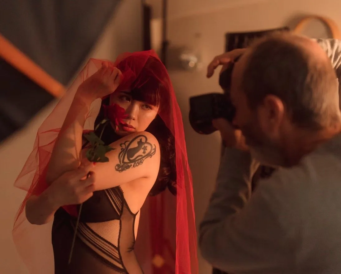 A woman with short red hair and tattoos posing for a photoshoot while wearing a black mesh outfit and a red veil, held by a photographer in a dimly lit room.