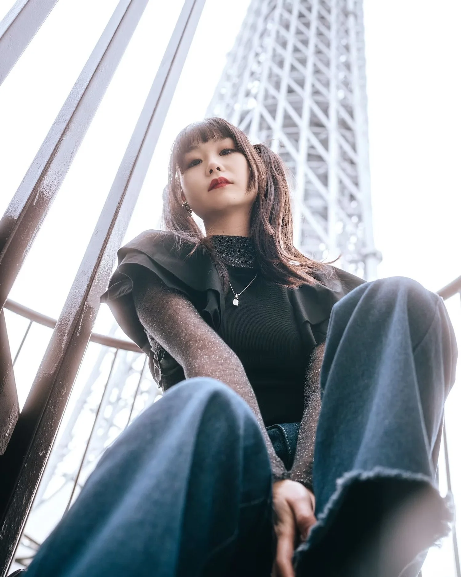 A young woman with brown hair styled in pigtails, wearing a black turtleneck with glittery sleeves and dark jeans, sitting outdoors near a metal fence with a tower in the background, looking down at the camera with a neutral expression.