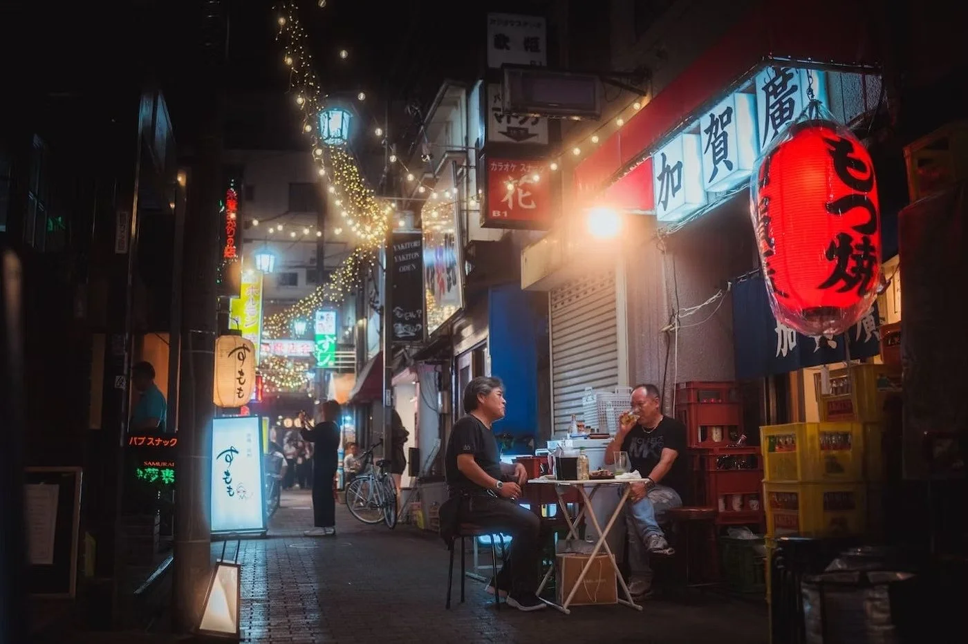 Nighttime street scene in Nakano, Tokyo Japan, with two men sitting at a small table outside a restaurant, illuminated by hanging string lights, paper lanterns, and neon signs.