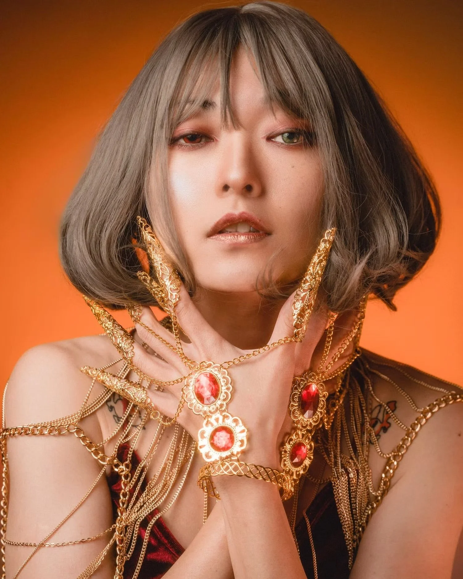 A young woman with short, wavy, grayish-brown hair and colorful contact lenses, wearing elaborate gold jewelry with red gemstones, reaching towards her face with her hands against an orange background.
