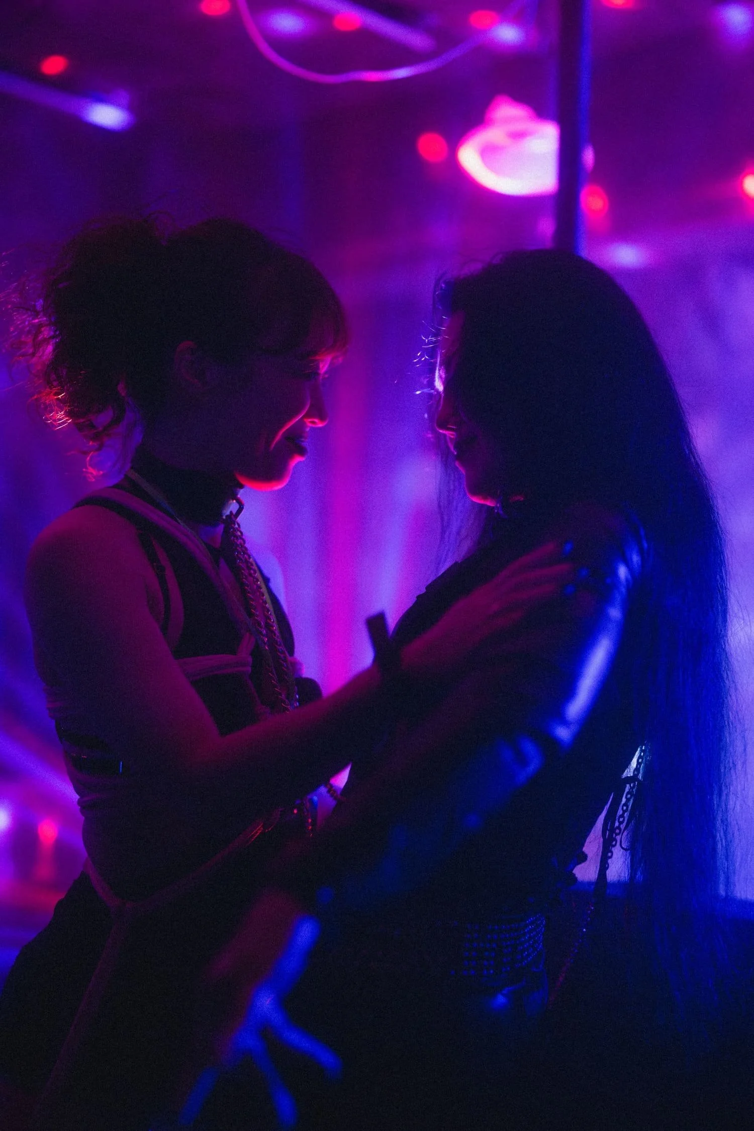 Bondage Ball 
---
Two women are facing each other in a dark, neon-lit environment with purple and pink lighting. The woman on the left has her hair tied back and is smiling, while the woman on the right has long hair and is holding the other woman. T