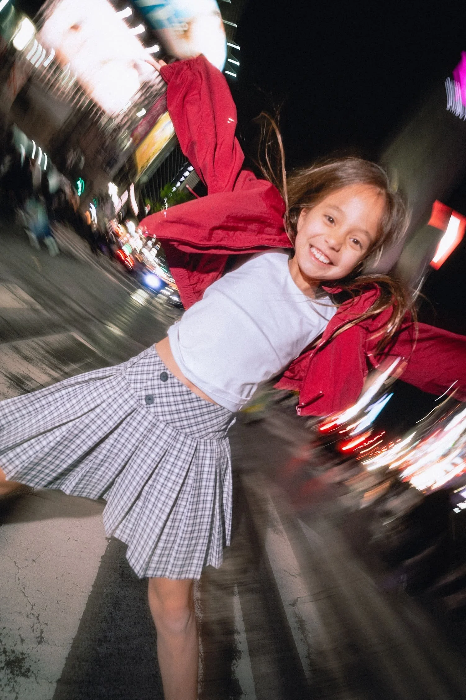 A smiling young girl with brown hair, wearing a plaid skirt, white shirt, and a red jacket, standing on a street at night with bright city lights of Shibuya crossing in the background.