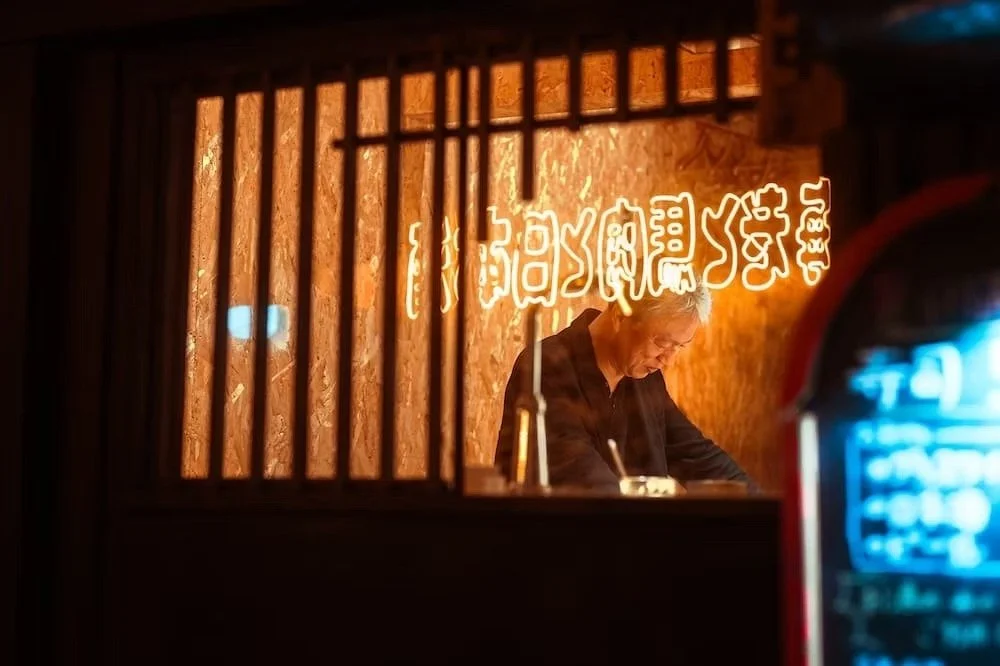 A man with gray hair in a dark shirt working inside a restaurant kitchen seen through a wooden window frame with vertical bars. Neon Chinese characters glow on the textured wall behind him. Ikebukuro neons by Tokyo street photographer Deniz Demir.