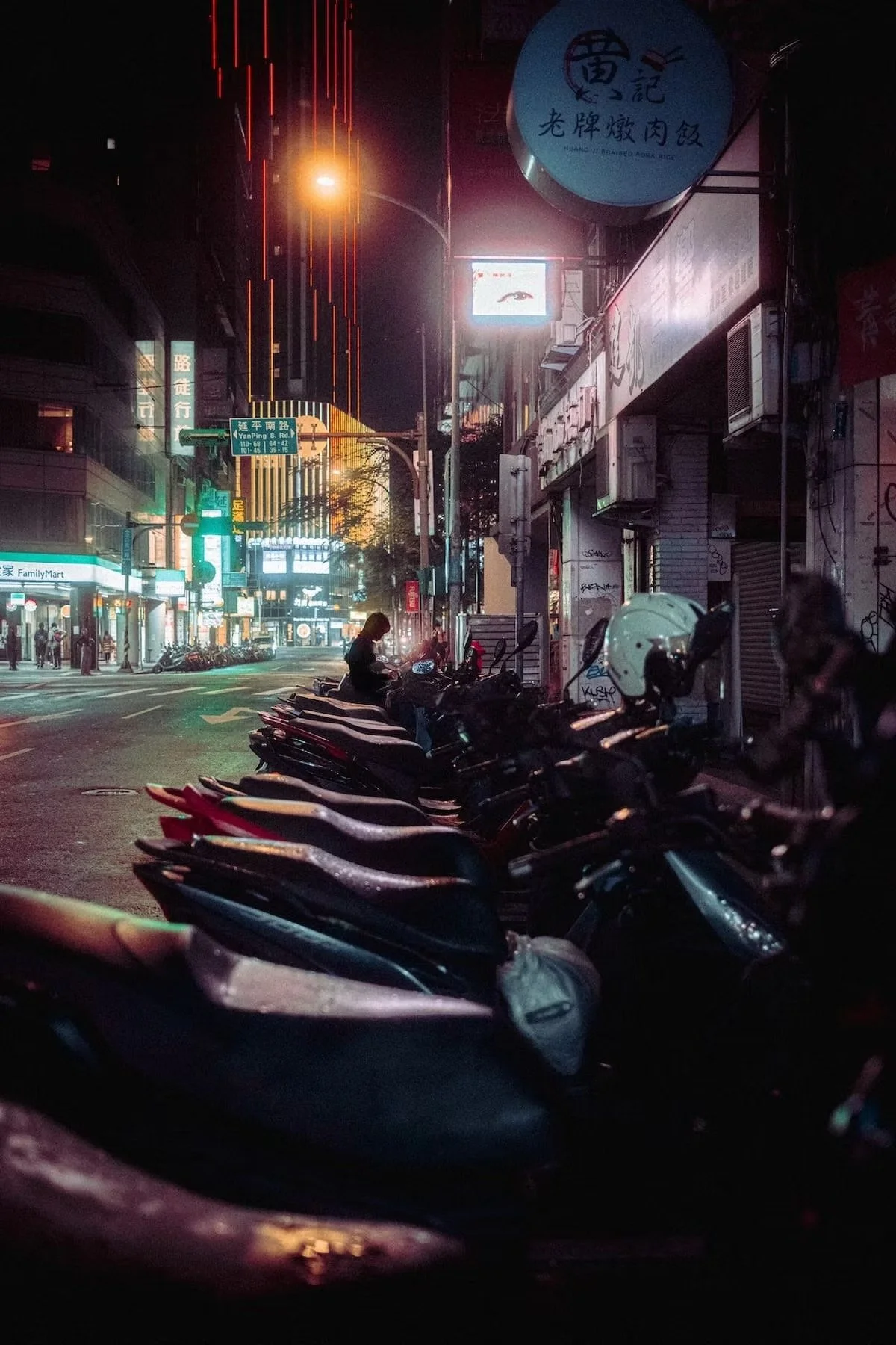 Nighttime city street scene with parked motorcycles in the foreground, illuminated signs, and a few pedestrians, including a woman looking at her phone, along the sidewalk. Neon lights and building signs are visible. Taipei night street photography b