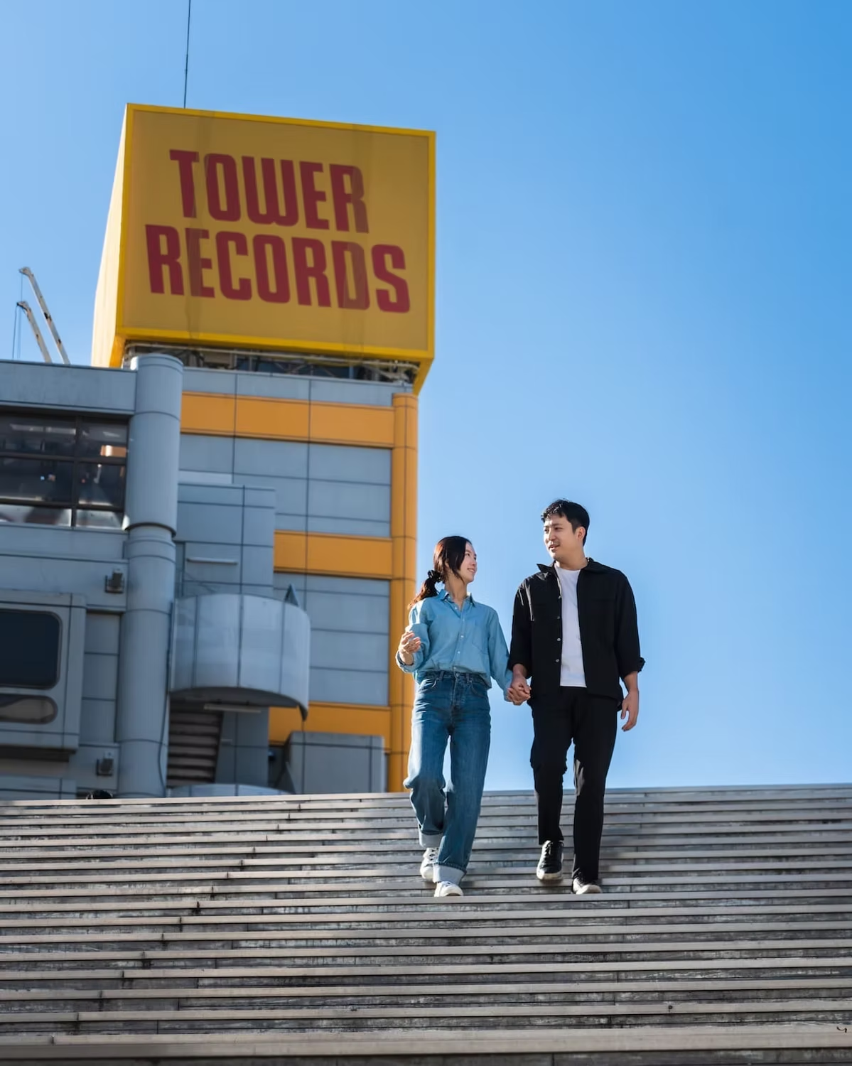 A couple walks down a set of wide outdoor stairs, holding hands. The scene is set in bright daylight, with the couple dressed casually in denim outfits against the urban backdrop.