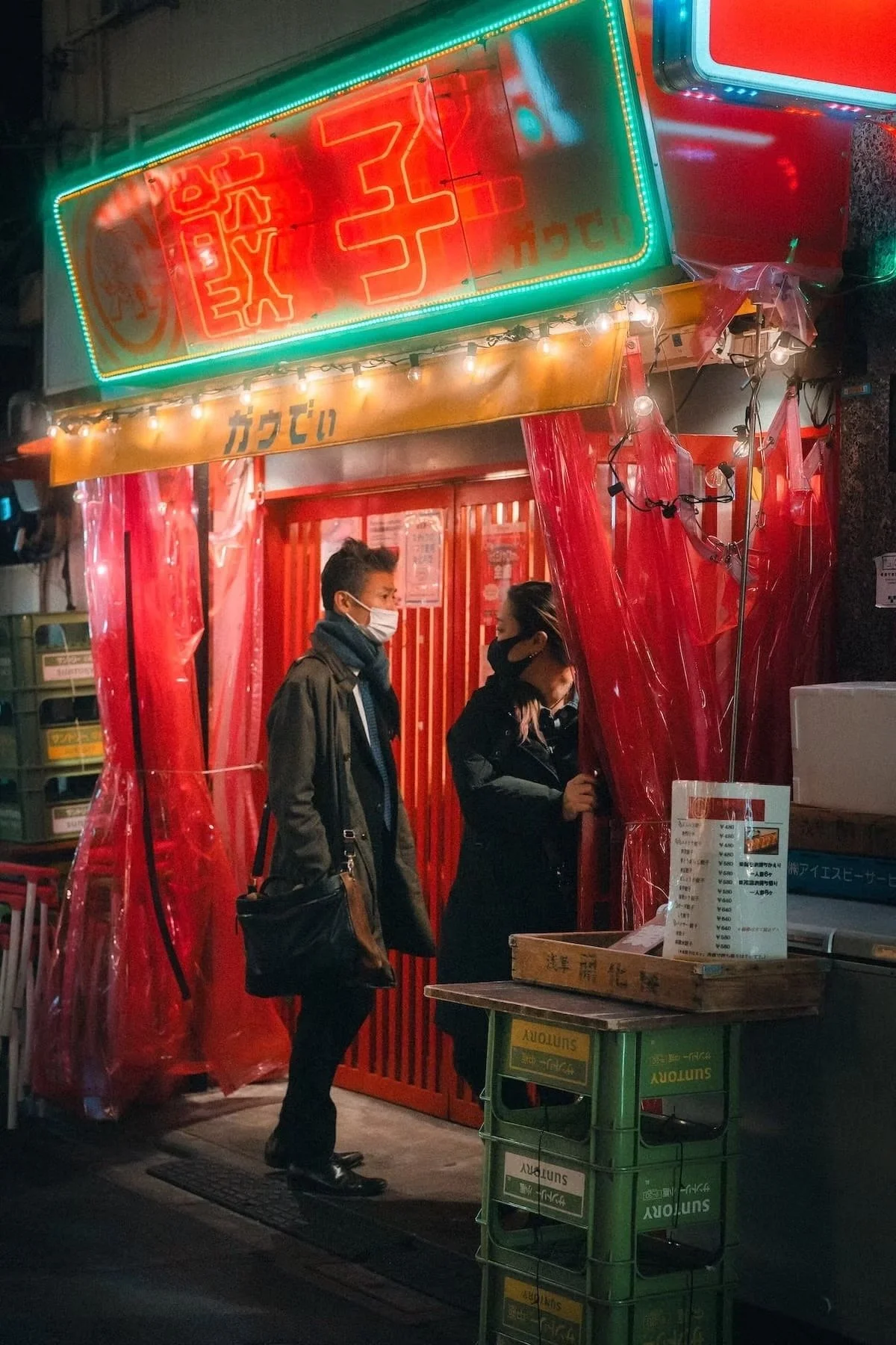 A brightly lit street food stall in Japan with red decorations, neon signage, and two people wearing masks standing inside, engaging in conversation.