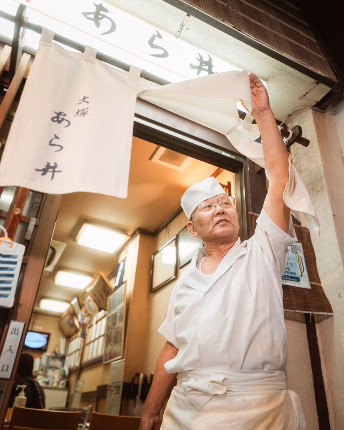 An elderly Japanese chef adjusting a noren curtain outside a restaurant. Shot by Tokyo street photographer Deniz Demir.