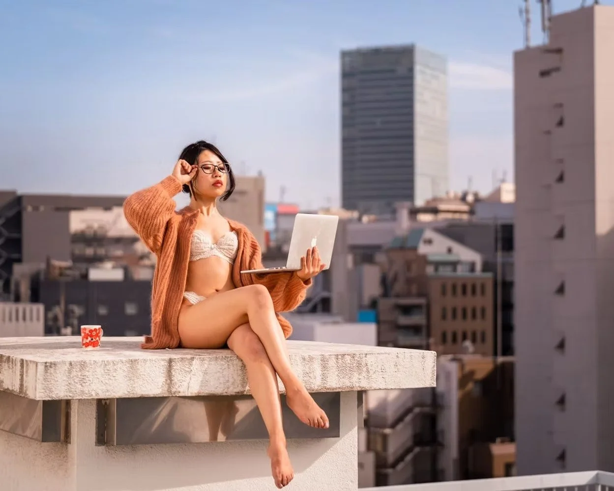 A woman sitting on a rooftop ledge in lingerie and a sweater, holding a laptop while adjusting her glasses with city buildings in the background. Tokyo rooftop