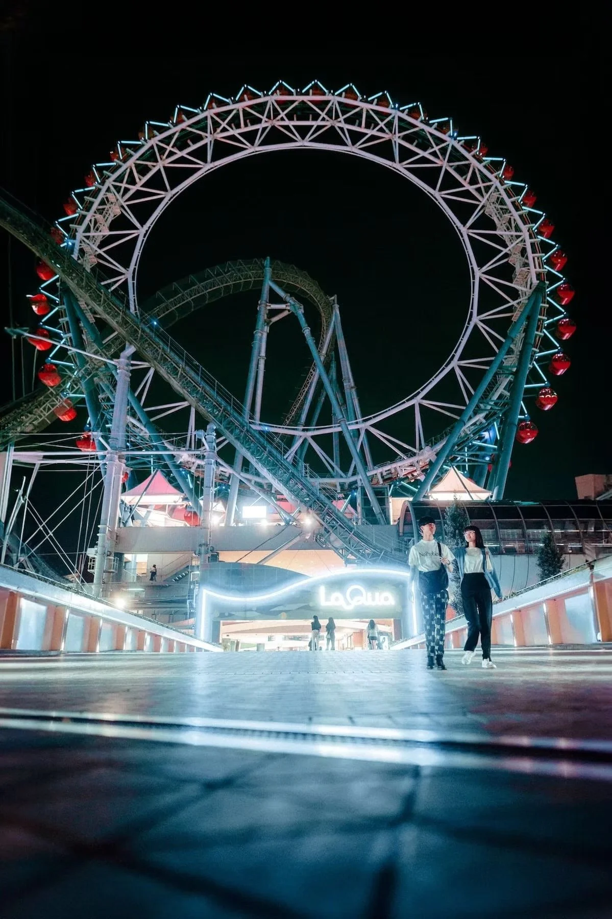 Night view of a lit-up Ferris wheel in Tokyo and roller coaster at an amusement park, with two women walking and talking in the foreground.