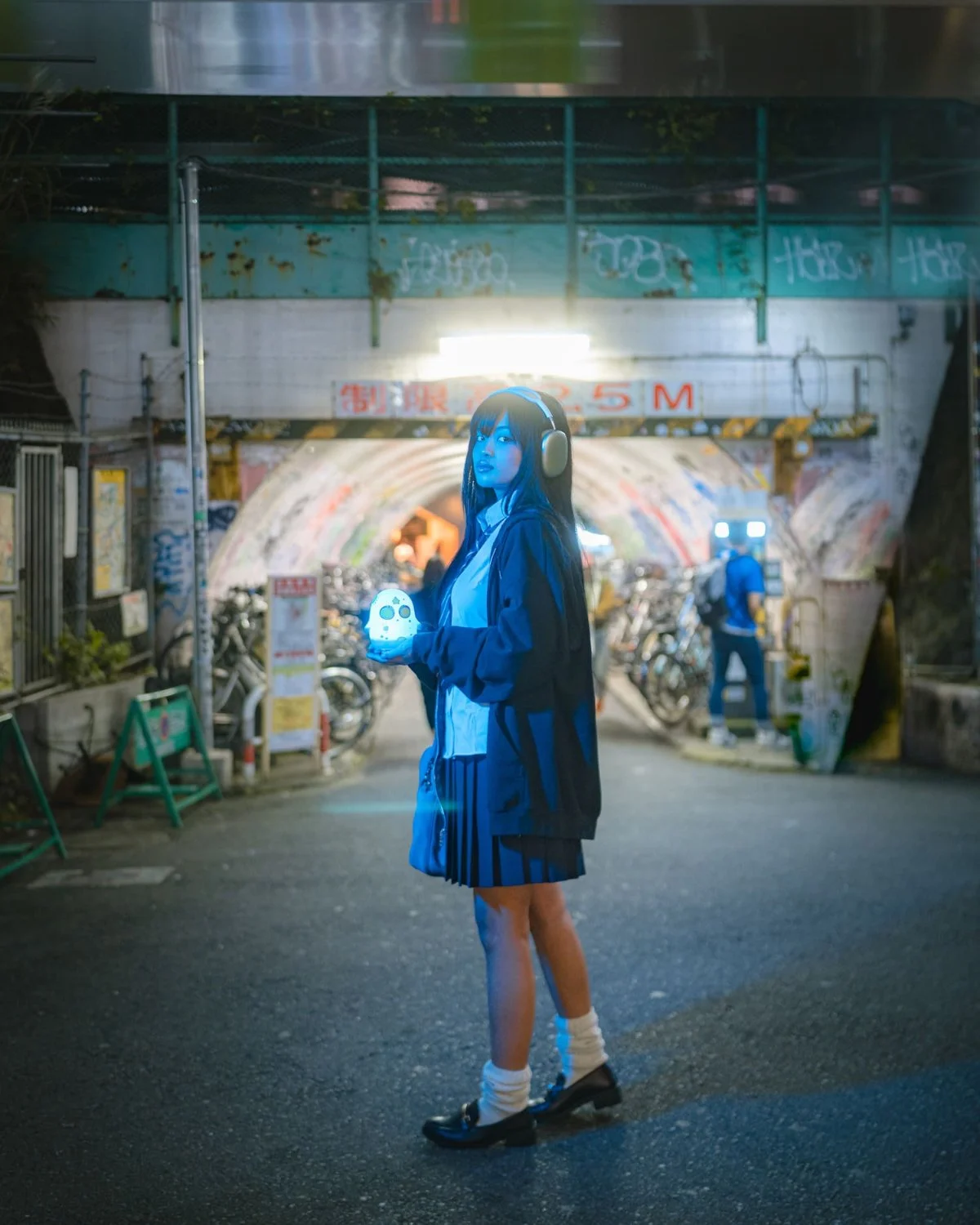 Young woman in school uniform and headphones standing under an underpass at night, holding a glowing object with a skull face, with bicycles and a person in the background.