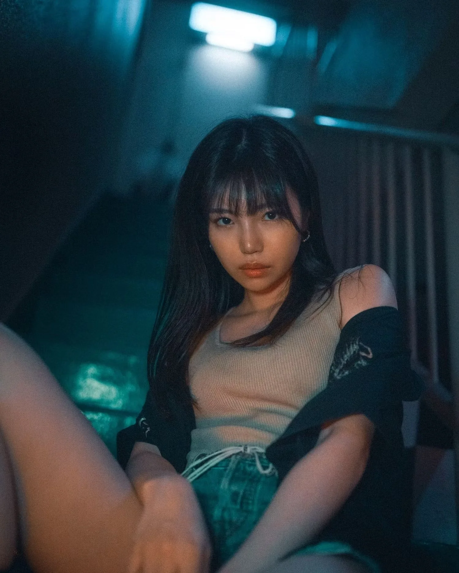 A young woman with long dark hair and bangs, wearing a beige top, sitting in a dimly lit room with a serious expression.