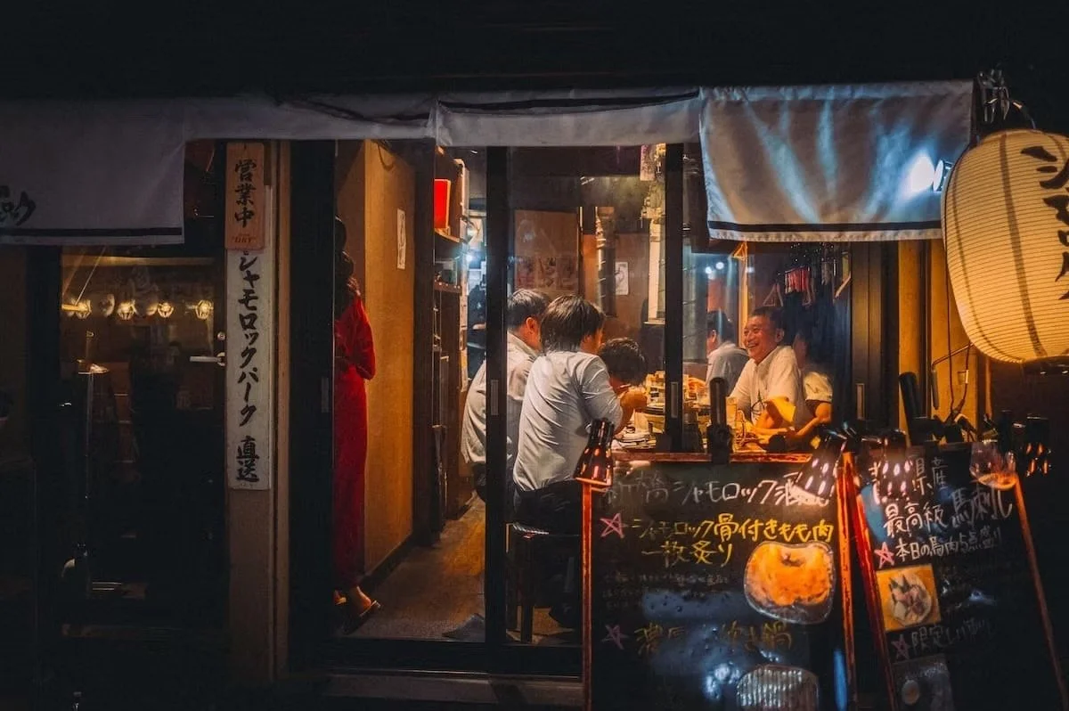 People dining inside a Japanese restaurant at night, with warm lighting, signs, and lanterns outside. Shimbashi Night scenes by Deniz Demir.