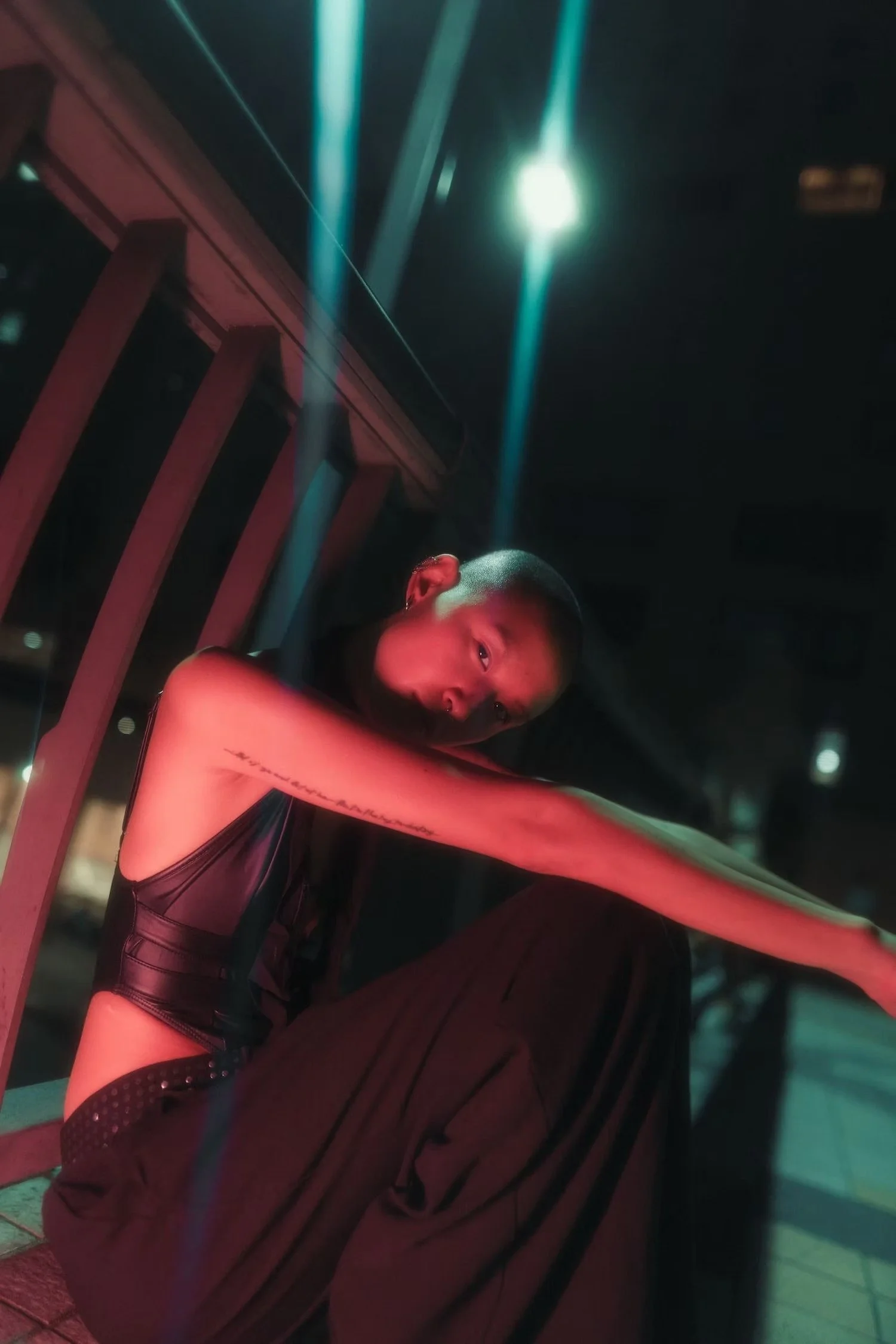 A young woman with short hair and earrings, dressed in a black outfit, sitting on a balcony at night with city lights in the background, illuminated by a nearby bright light.