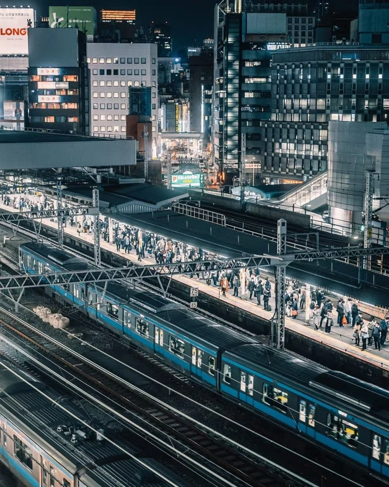 Night view of a busy train station in a city with many illuminated office buildings and skyscrapers, crowded platform with people waiting, and trains on multiple tracks.