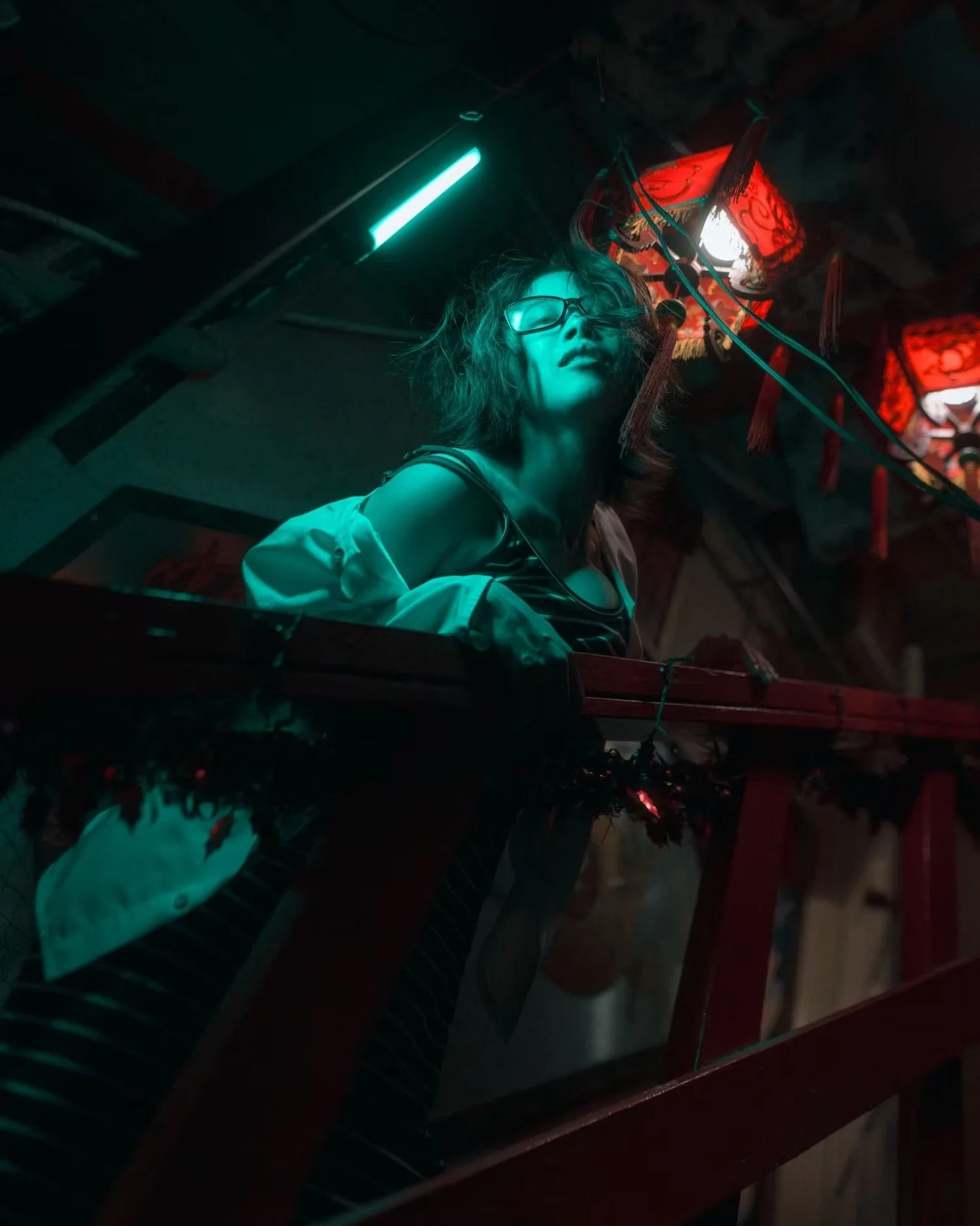 A woman with wet hair, glasses, and a striped shirt looking upward, illuminated by red and green neon lights, standing behind a glass display case filled with stuffed animals, in a dimly-lit room with hanging red paper lanterns.