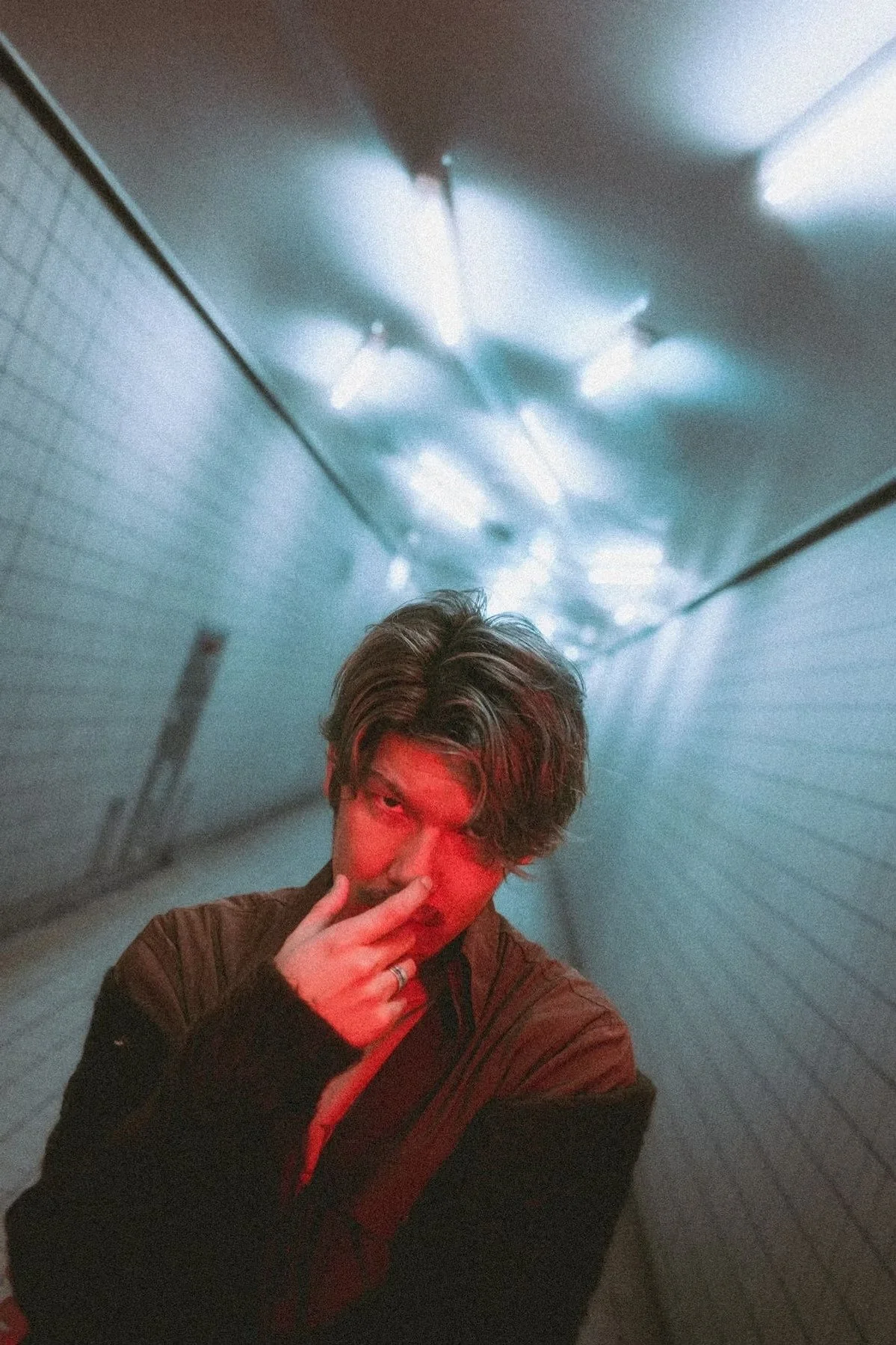 A Japanese model with wavy brown hair standing in a dimly lit tunnel with blue lighting, making a peace sign near their face. Shot by Tokyo portrait photographer Deniz Demir in a train station.