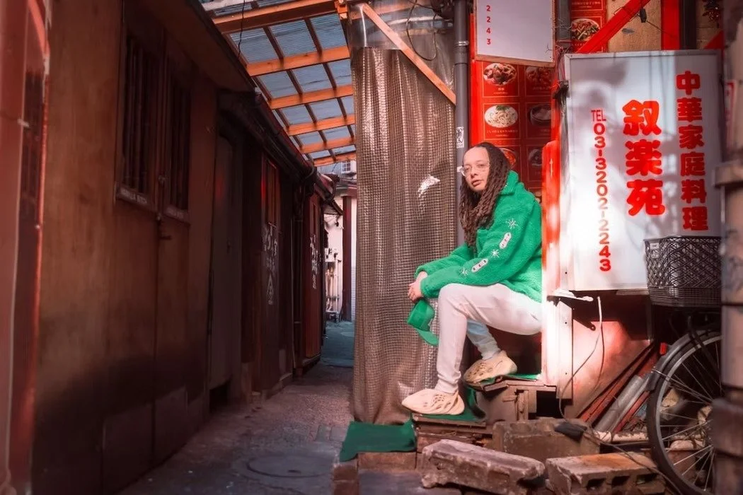 A woman with glasses and long dreadlocks sitting on a small ledge in an alleyway, wearing a green hoodie, white pants, and beige shoes, surrounded by red signs and structures in an urban setting.