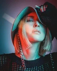 A woman wearing a wide-brimmed hat, holding the hat with her gloved hand, with a short blonde bob haircut, in a posed portrait with colorful lighting.