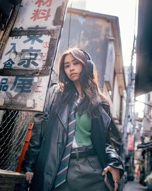 A young woman with long wavy hair wearing headphones, a green sweater, a school tie, and a black leather jacket, standing on a busy city street.