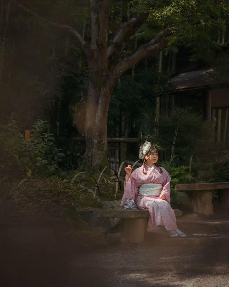 A woman dressed in a pink kimono sits on a wooden bench in a serene, wooded outdoor area with trees and greenery surrounding her.