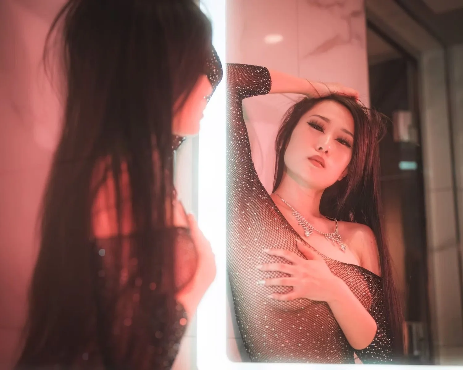 A woman with long dark hair posing in front of a mirror, wearing a sparkly black dress and jewelry, with her hand in her hair and looking at herself.