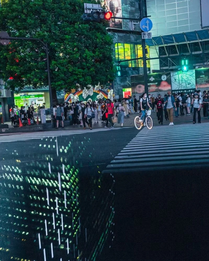 A busy city crosswalk filled with pedestrians, a cyclist on the crosswalk, traffic lights showing green and red, digital billboards, and a large building with reflective glass windows in the background.