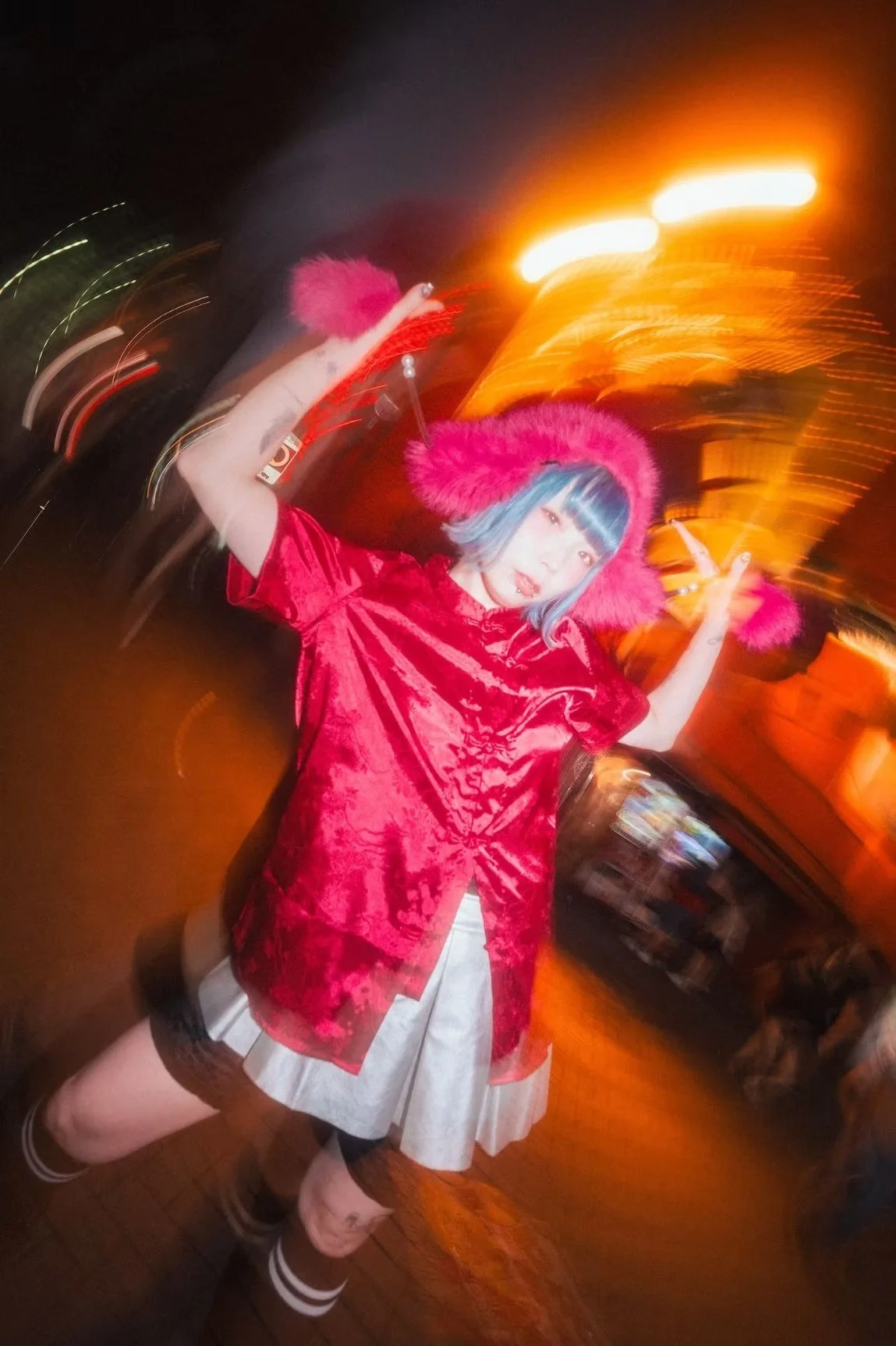 A young woman with blue hair and a red outfit strikes a playful pose on a busy Tokyo street at night. Bright orange lights and a motion blur effect capture the energy and excitement of the city. Photography by Deniz Demir.