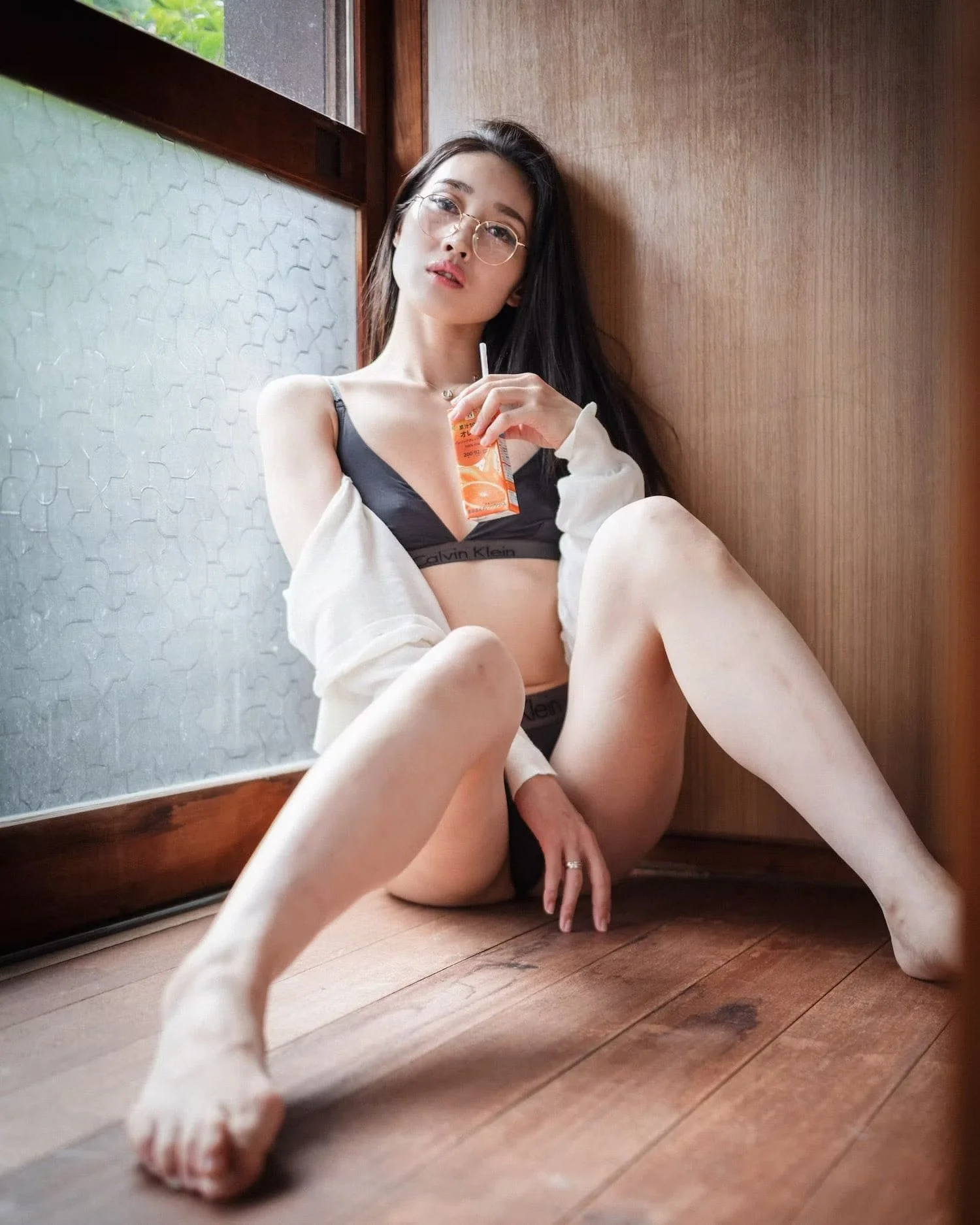 A young woman with long dark hair, glasses, and minimal makeup sitting on a wooden floor next to a textured glass window and wooden wall, holding an orange juice box.