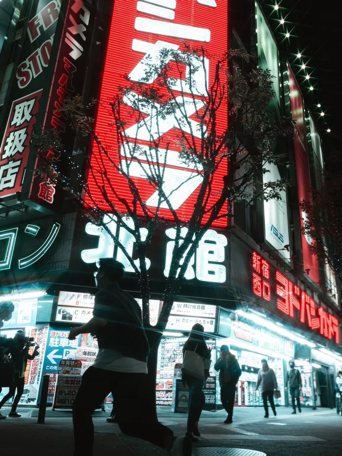 Night scene of a busy street in Japan with illuminated signs in Japanese, a large red and white neon advertisement, people walking, and a leafless tree in the foreground.
