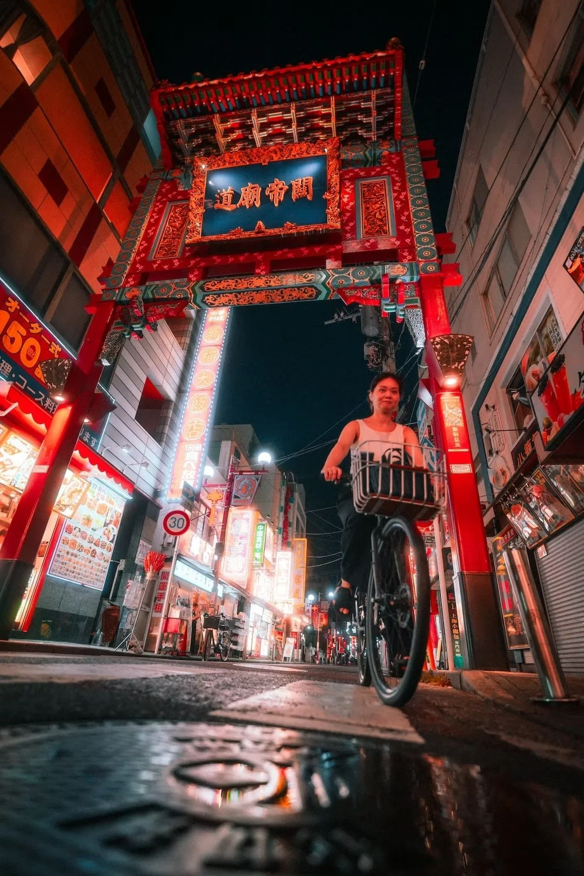 A woman riding a bicycle through a brightly lit Chinatown street at night in Yokohama Japan. The street is decorated with colorful signs and neon lights, with a traditional Chinese gate overhead.