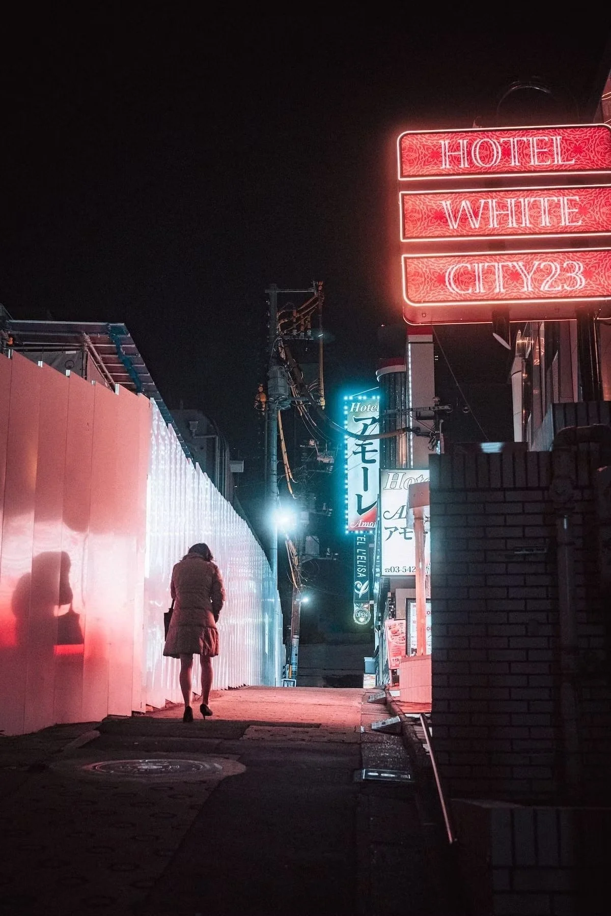 A person walking on a city sidewalk at night, illuminated by neon signs including one that reads 'HOTEL WHITE CITY23' in red. The scene features various lit signs and power lines along the street in Shibuya Tokyo.