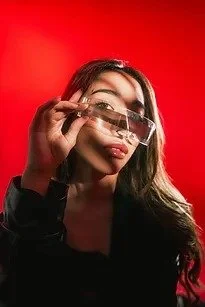 Person with long dark hair holding sunglasses against a red background