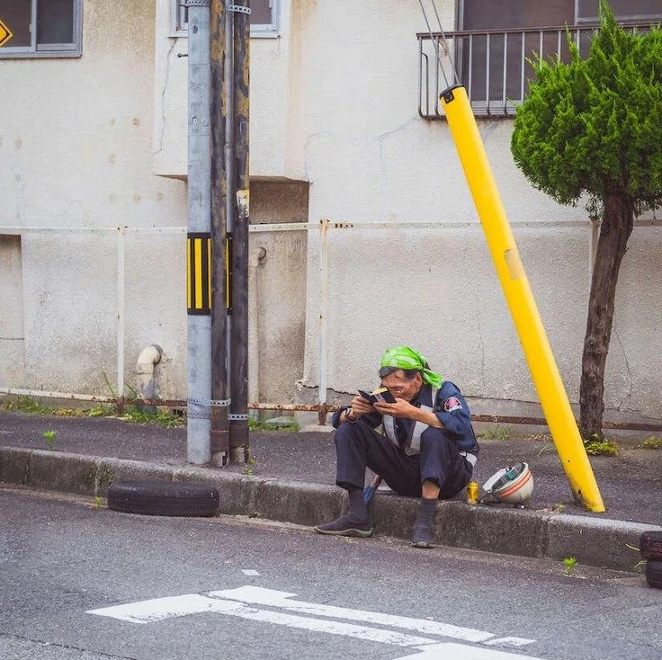 A man sitting on the street in Osaka with a yellow pole nearby, captured during slow travel.