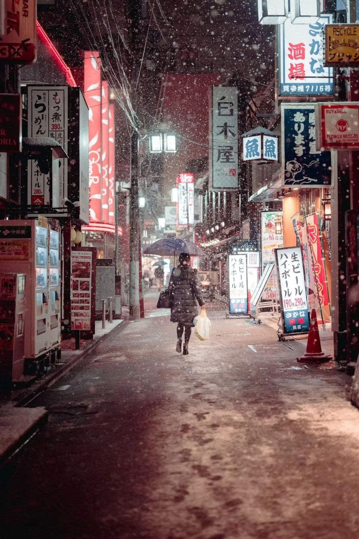 Snowy street scene at night with illuminated signs in Japanese, a person walking with an umbrella and carrying shopping bags, snow falling and creating a misty atmosphere in Nakano shot by Tokyo street photographer Deniz Demir.