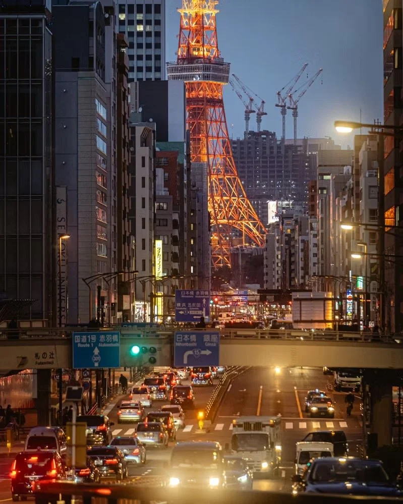 A busy city street at dusk with numerous cars and traffic lights, with Tokyo Tower illuminated in the background and cranes visible in a construction site.