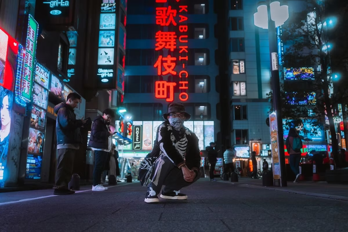A person dressed in dark attire crouches in front of neon lights in Tokyo's Kabukicho entertainment district at night.