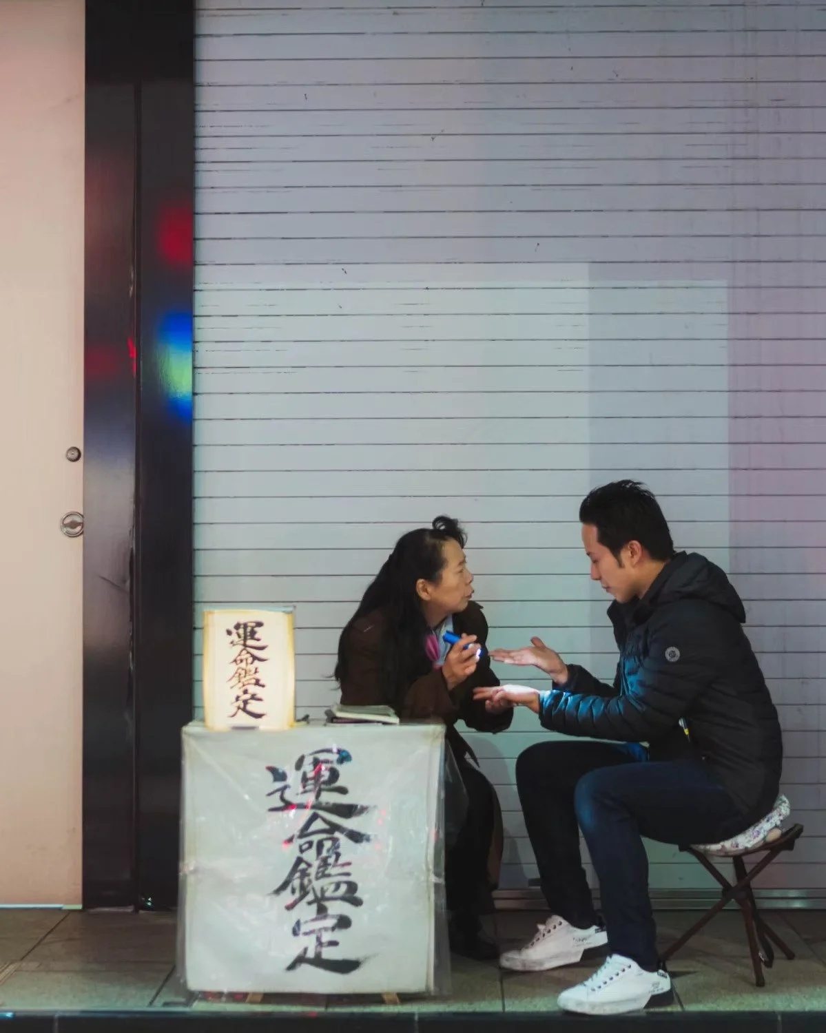 A woman and a man sitting and talking in front of a shop with closed shutters. The woman appears to be explaining something, holding a small object in her hand. The man is listening attentively, leaning forward with his palms open. There is a sign wi