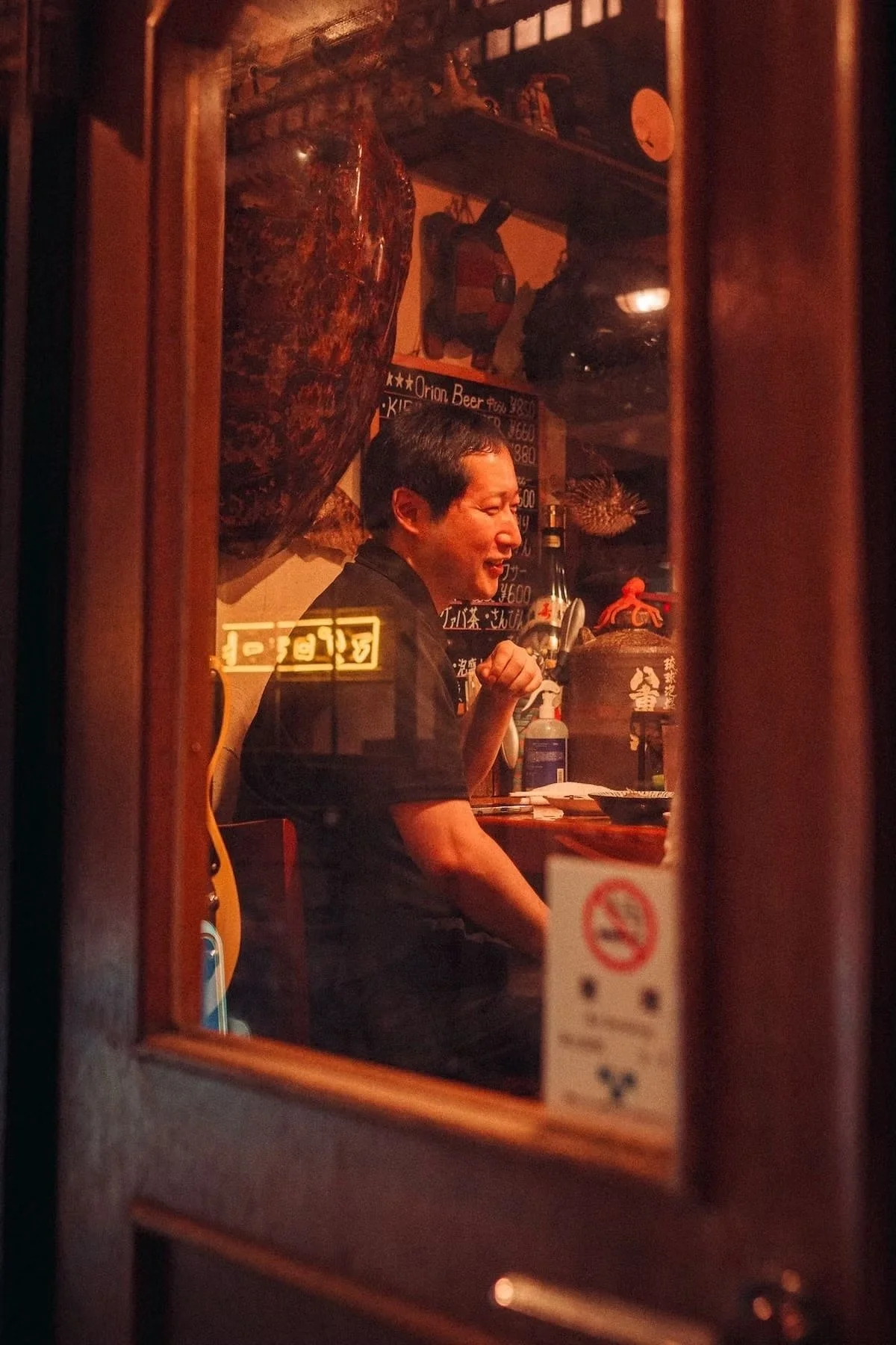 A man sitting inside a Japanese restaurant, seen through a window. The man appears to be smiling or laughing, with various items and decorations, including a chalkboard menu, bottles, and traditional Japanese decor, visible in the background.