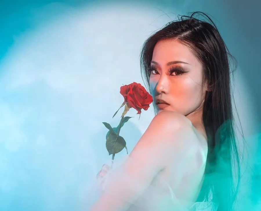 A woman poses with a rose, bathed in soft blue and green light. Her expression is calm yet captivating, creating an elegant and serene atmosphere.