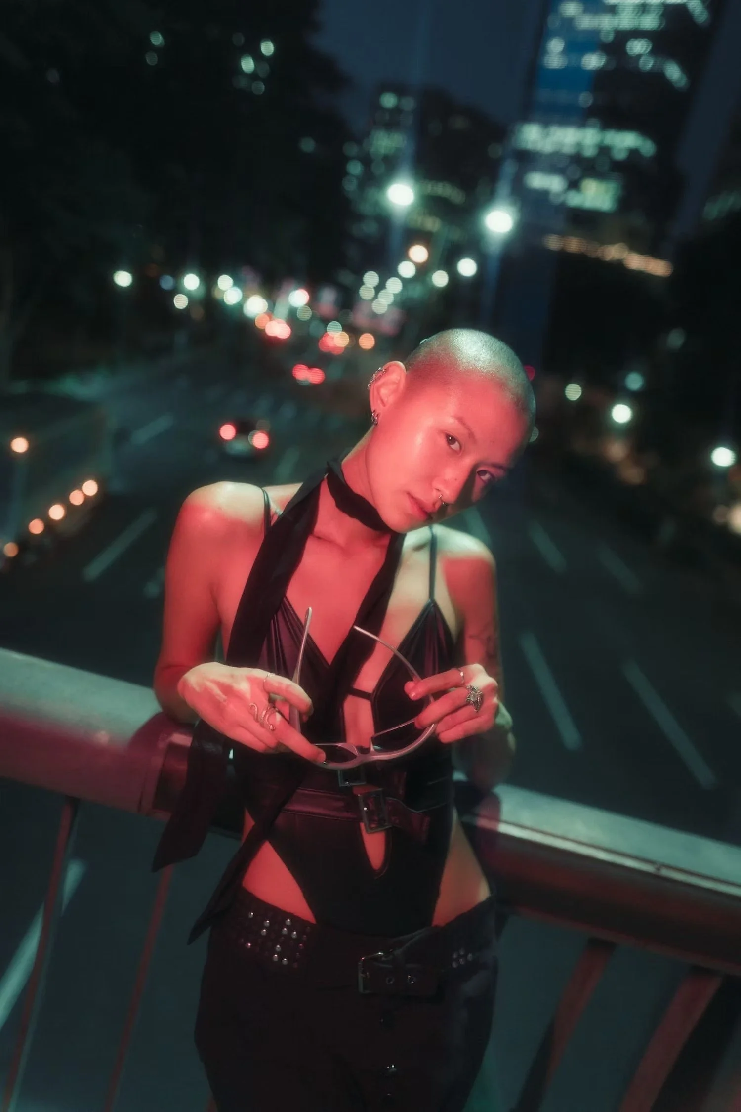 A person with a shaved head and earrings posing inside an elevator with city lights and buildings visible through the window behind them.