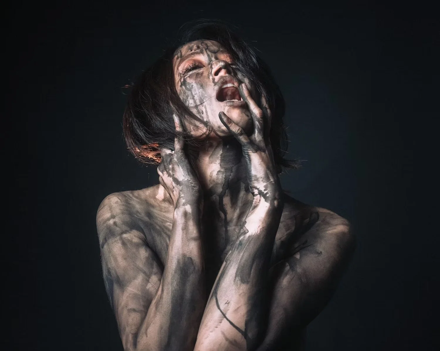 A person with black hair has their head tilted back and eyes closed, with hands touching their face, covered in black ink or paint against a dark background.