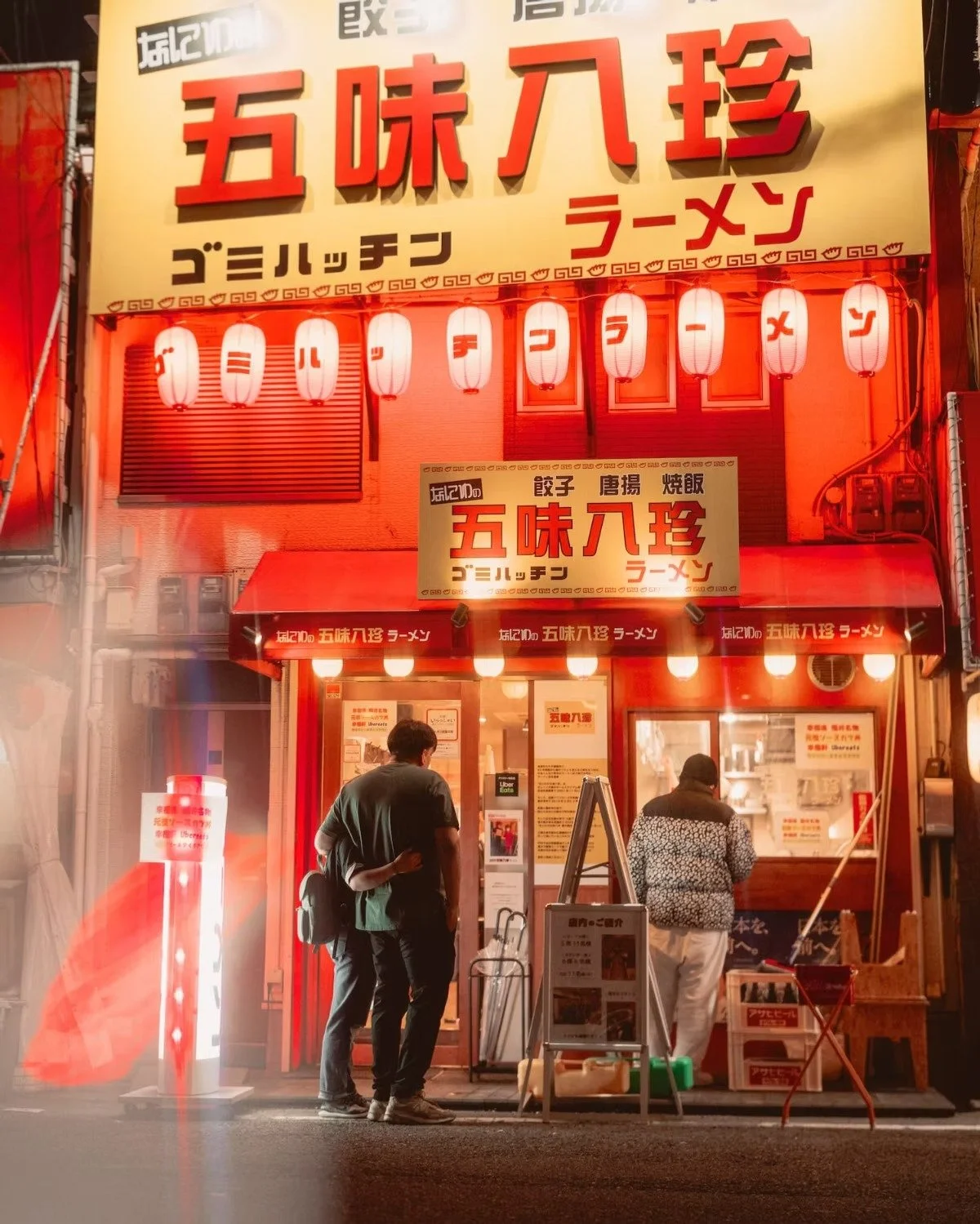 Nighttime view of a Japanese ramen restaurant decorated with red lanterns and large red and yellow signs with Japanese characters and some English words. Two people are standing outside the entrance, looking at the menu or ordering. Osaka Dotonbori.