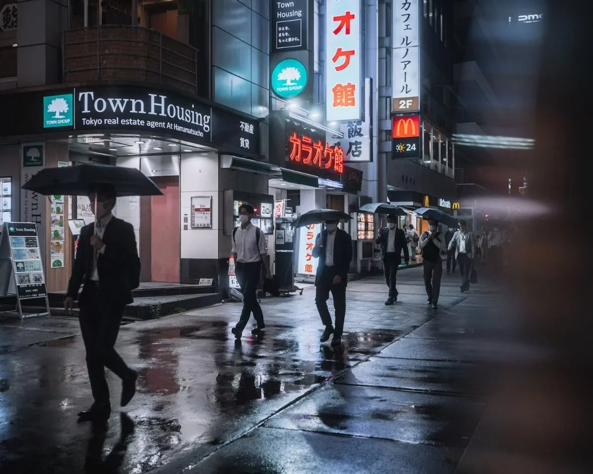 City street scene at night with people walking under umbrellas in the rain. Signs for Town Housing, a karaoke bar, and McDonald's are visible on the buildings. Night street photography.