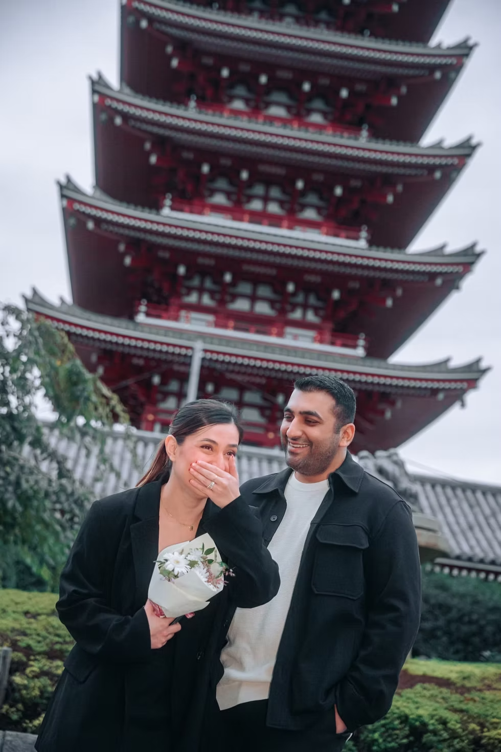 A woman and a man smiling together in front of a traditional Japanese pagoda, with the woman holding a small bouquet of flowers.