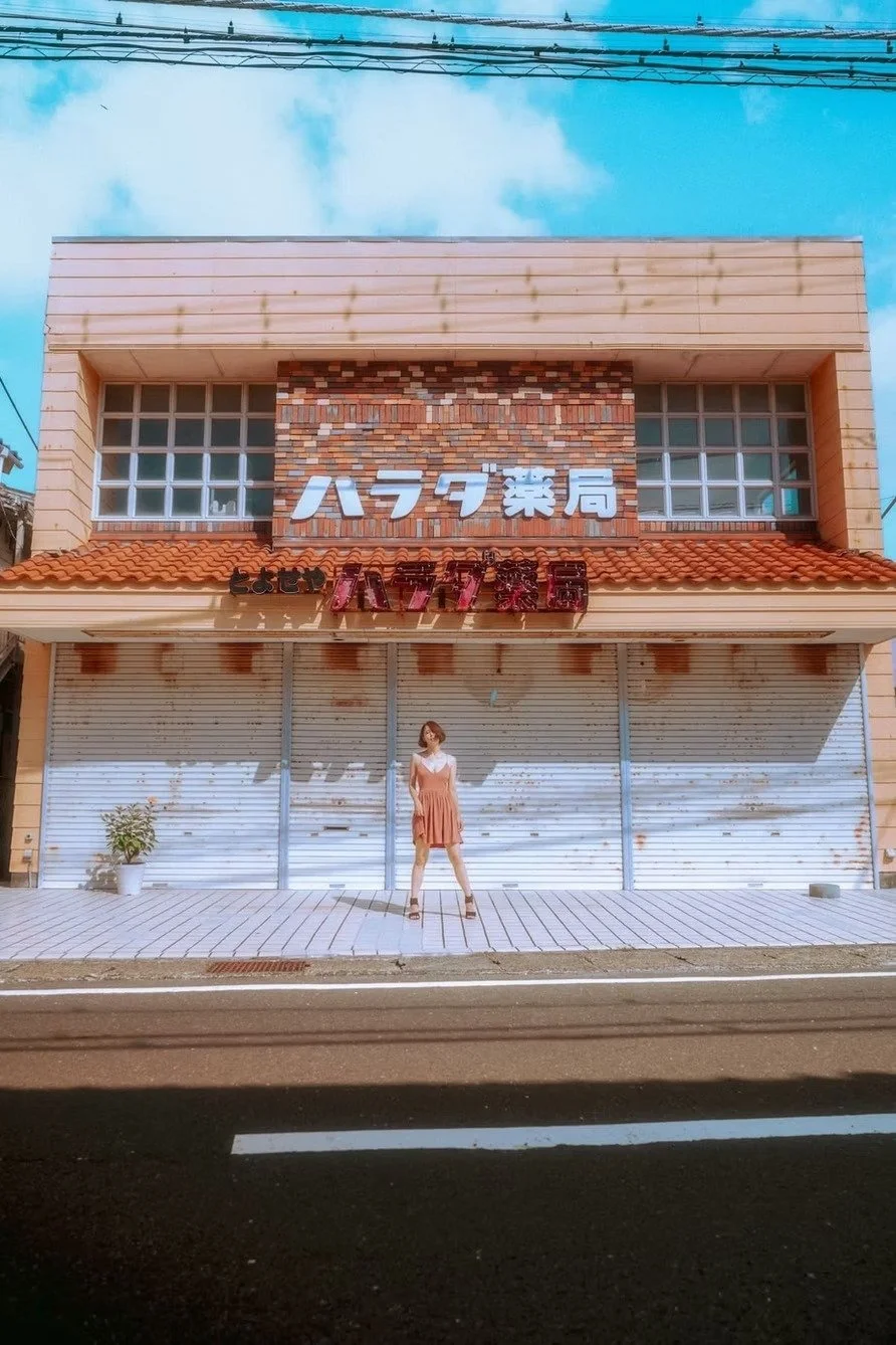A woman in a pink dress and sandals standing in front of a closed storefront with Japanese signs. The building has a brick and beige exterior with a red-tiled roof, and there is a potted plant to the left.
