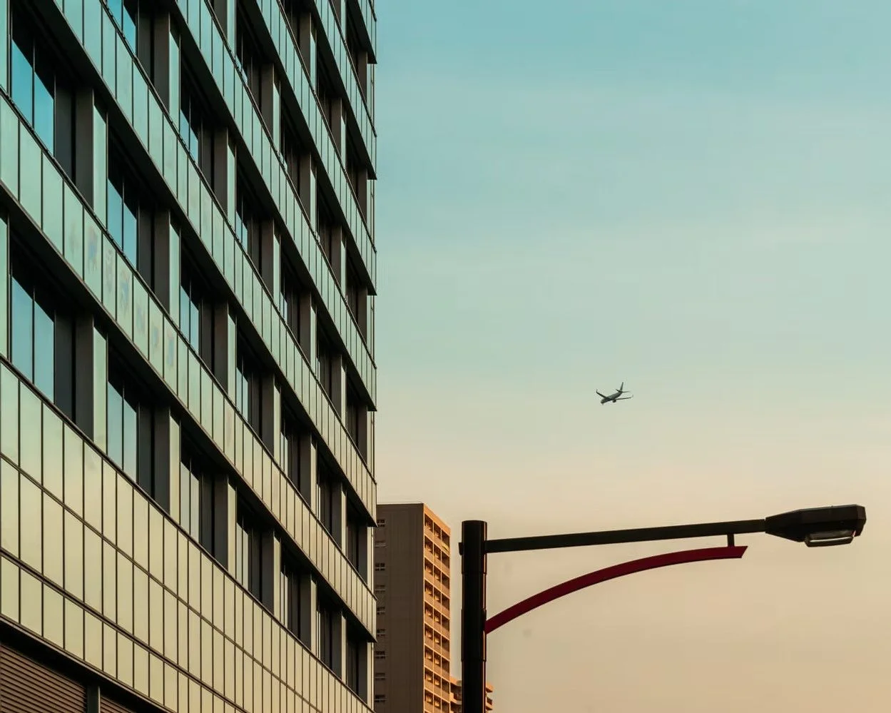 Cityscape with a tall glass building on the left, a streetlamp in the foreground, and a plane flying in the sky during sunset or sunrise. Captured by Deniz Demir.