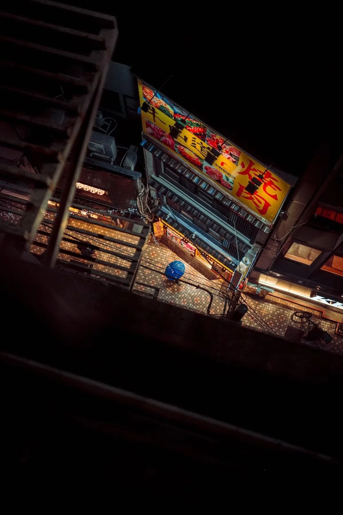 Nighttime view of a narrow, multi-story building with bright, colorful signs with Japanese kanji and a small street below with tiles and a blue umbrella. Nakano Tokyo from above