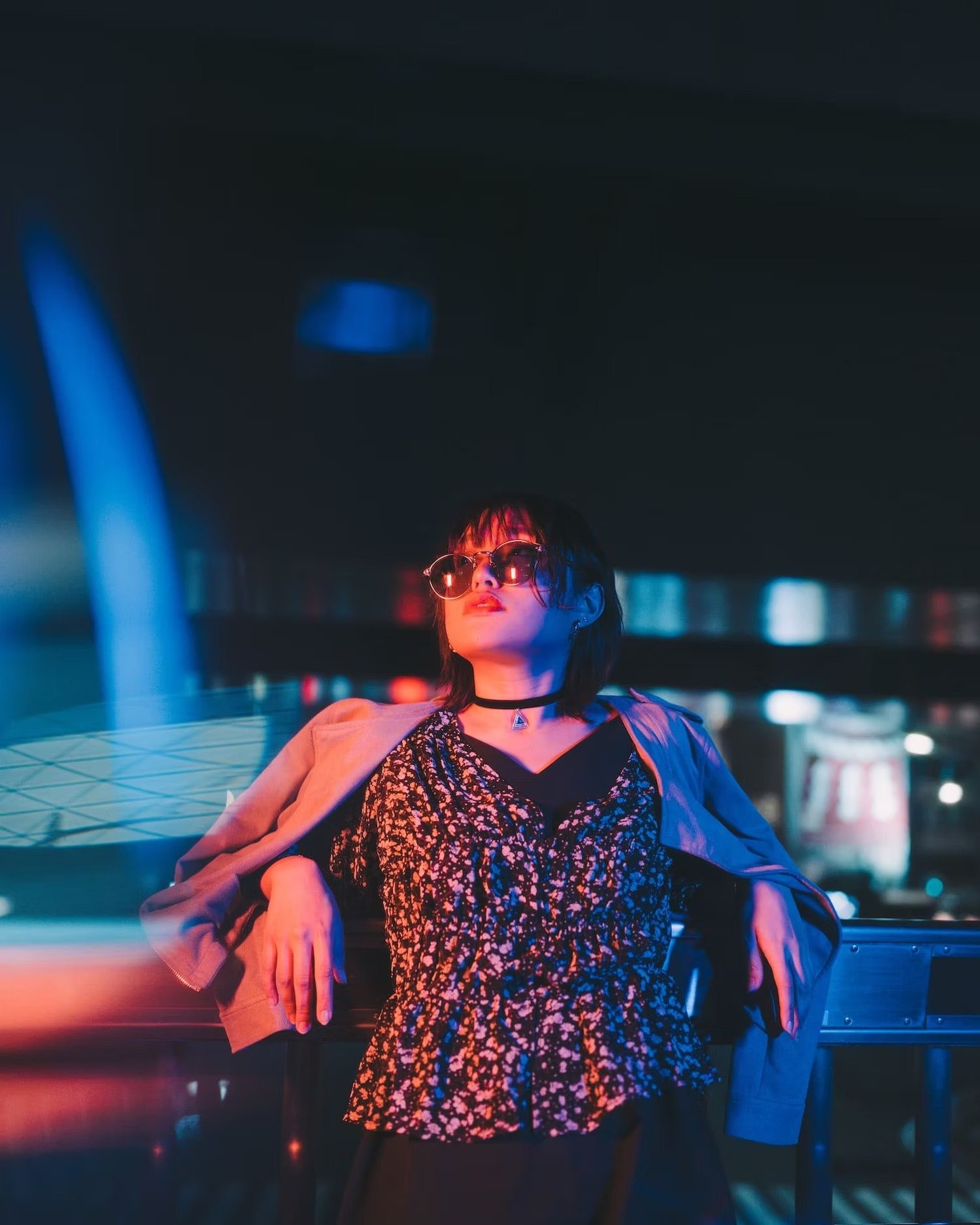 A woman with short dark hair and sunglasses sitting on a bench in a city at night, illuminated by colorful neon lights.