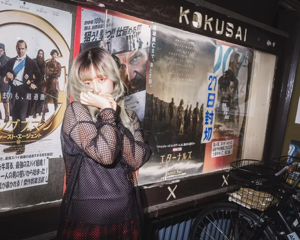A woman with dyed blonde hair covering part of her face with her hand, standing in front of a Japanese movie poster display outside a movie theater called 'KOKUSAI'. She is wearing a black fishnet dress.